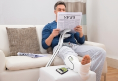 Disabled Man With Crutches Sitting On Sofa Reading Newspaper